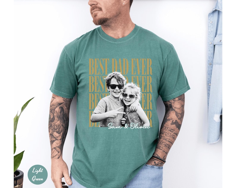 May include: Light green t-shirt with the text "BEST DAD EVER" in gold lettering. A black and white photo of two children wearing sunglasses is printed on the shirt, with the names "Sam & Olivia" below.