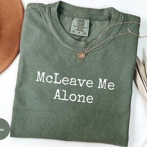 K&ouml;nnte beinhalten: Ein moosgr&uuml;nes T-Shirt mit dem wei&szlig;en Text "McLeave Me Alone". Das Shirt hat einen Rundhalsausschnitt und kurze &Auml;rmel. Eine silberne Halskette mit einem kleinen Anh&auml;nger liegt &uuml;ber dem Shirt.