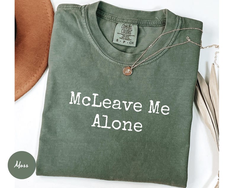 K&ouml;nnte beinhalten: Ein moosgr&uuml;nes T-Shirt mit dem wei&szlig;en Text "McLeave Me Alone". Das Shirt hat einen Rundhalsausschnitt und kurze &Auml;rmel. Eine silberne Halskette mit einem kleinen Anh&auml;nger liegt &uuml;ber dem Shirt.