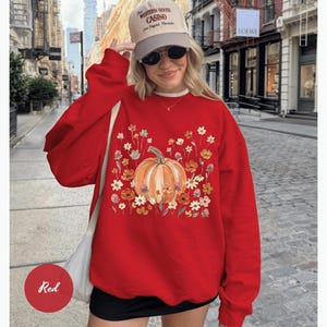 May include: A red crewneck sweatshirt featuring a central graphic of an orange pumpkin surrounded by a variety of colorful autumn flowers in shades of yellow, orange, and white. The sweatshirt has long sleeves and a relaxed fit.