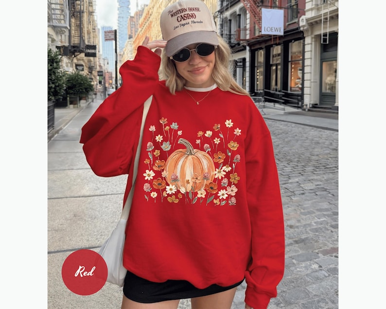 May include: A red crewneck sweatshirt featuring a central graphic of an orange pumpkin surrounded by a variety of colorful autumn flowers in shades of yellow, orange, and white. The sweatshirt has long sleeves and a relaxed fit.