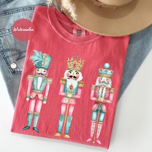 May include: A red t-shirt featuring three colorful nutcracker illustrations. The nutcrackers are depicted in pink, teal, and gold, with ornate details. The shirt is folded on top of blue jeans, with a hat and necklace visible.