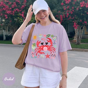 May include: A light purple t-shirt featuring a red crab design with hearts, cocktails, and seashells. The shirt is paired with white shorts and a white baseball cap. A brown shoulder bag is also visible.