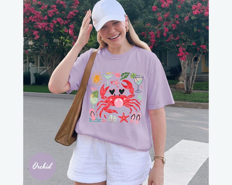 May include: A light purple t-shirt featuring a red crab design with hearts, cocktails, and seashells. The shirt is paired with white shorts and a white baseball cap. A brown shoulder bag is also visible.