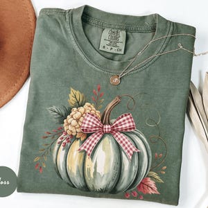 May include: A moss green t-shirt with a watercolour pumpkin design and a red and white checked bow. The pumpkin is surrounded by autumn leaves. A gold necklace rests on the shirt. The shirt has a Comfort Colors tag.