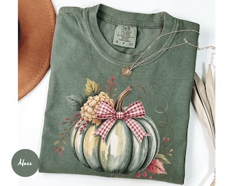 May include: A moss green t-shirt featuring a watercolor pumpkin design with a red and white checkered bow. The pumpkin is surrounded by fall foliage. A gold necklace rests on the shirt. The shirt has a Comfort Colors tag.