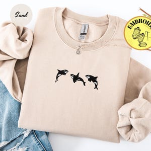 May include: A sand-colored sweatshirt with a crew neck and long sleeves. The sweatshirt features a small embroidered design of three black orcas. A silver necklace with a sun pendant is visible. The word "Embroidery" is in a yellow circle.