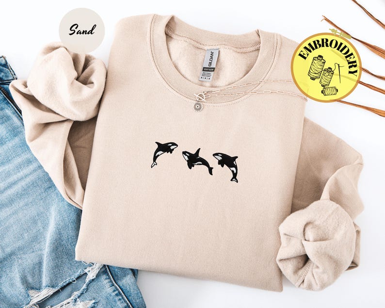 May include: A sand-colored sweatshirt with a crew neck and long sleeves. The sweatshirt features a small embroidered design of three black orcas. A silver necklace with a sun pendant is visible. The word "Embroidery" is in a yellow circle.