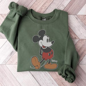 May include: An olive green sweatshirt featuring a graphic of Mickey Mouse in his classic pose. Mickey is depicted in black, red, and tan colors. The Disney logo is printed in white near Mickey's feet. The sweatshirt has cuffed sleeves.