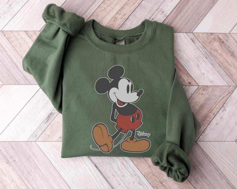 May include: An olive green sweatshirt featuring a graphic of Mickey Mouse in his classic pose. Mickey is depicted in black, red, and tan colors. The Disney logo is printed in white near Mickey's feet. The sweatshirt has cuffed sleeves.