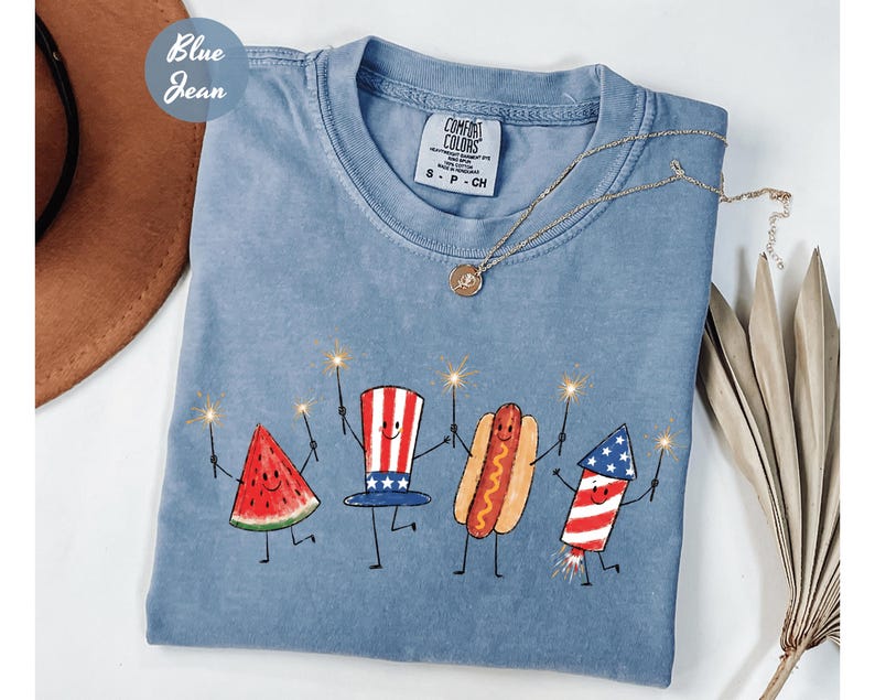 K&ouml;nnte beinhalten: Hellblaues Jeans-T-Shirt mit einem Motiv aus Wassermelone, Uncle Sam-Hut, Hotdog und Feuerwerk, die jeweils einen Wunderkerzenstab halten. Das Shirt hat einen Rundhalsausschnitt und kurze &Auml;rmel. Eine goldene Halskette mit Anh&auml;nger liegt &uuml;ber dem Shirt.