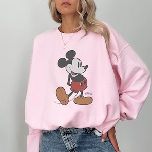 May include: A light pink sweatshirt featuring a classic Mickey Mouse graphic. The cartoon character is depicted in his iconic pose, wearing red shorts, brown shoes, and white gloves. The Disney logo is visible.