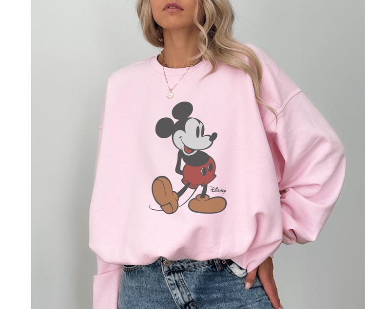 May include: A light pink sweatshirt featuring a classic Mickey Mouse graphic. The cartoon character is depicted in his iconic pose, wearing red shorts, brown shoes, and white gloves. The Disney logo is visible.