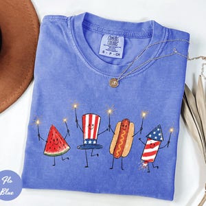 K&ouml;nnte beinhalten: Ein blaues Comfort Colors T-Shirt mit einer skurrilen Illustration einer Wassermelonenscheibe, einem Hut, einem Hotdog und einem Feuerwerk, die jeweils Wunderkerzen halten. Das Shirt hat einen Rundhalsausschnitt und ein kleines Etikett. Eine goldene Halskette liegt &uuml;ber dem Shirt.