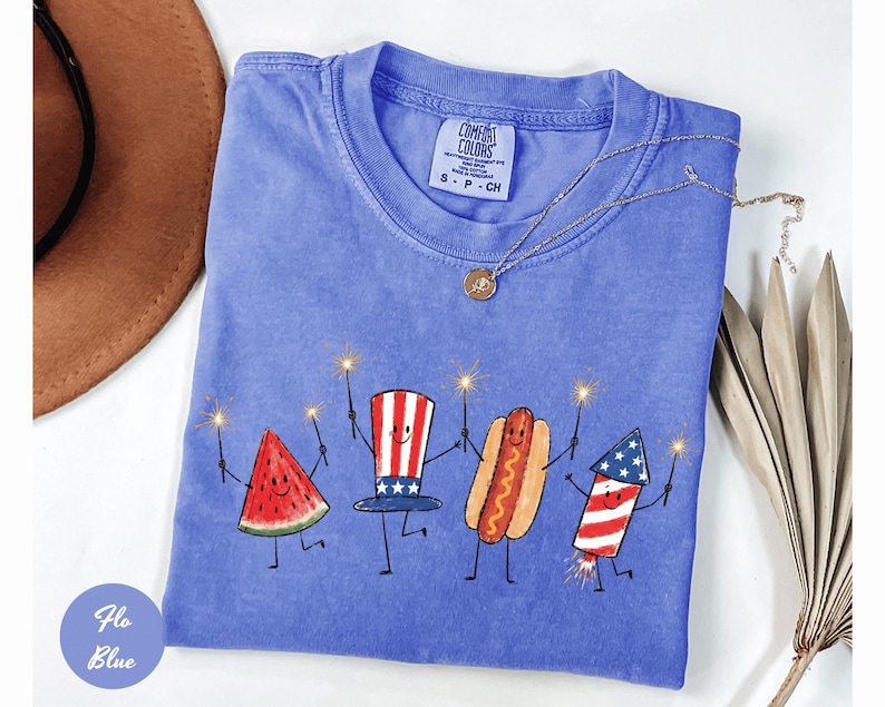 K&ouml;nnte beinhalten: Ein blaues Comfort Colors T-Shirt mit einer skurrilen Illustration einer Wassermelonenscheibe, einem Hut, einem Hotdog und einem Feuerwerk, die jeweils Wunderkerzen halten. Das Shirt hat einen Rundhalsausschnitt und ein kleines Etikett. Eine goldene Halskette liegt &uuml;ber dem Shirt.