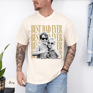 May include: Ivory-colored t-shirt with the text "BEST DAD EVER" in gold. A black and white photo of two children wearing sunglasses is centered on the shirt, with the names "Sam & Olivia" below.
