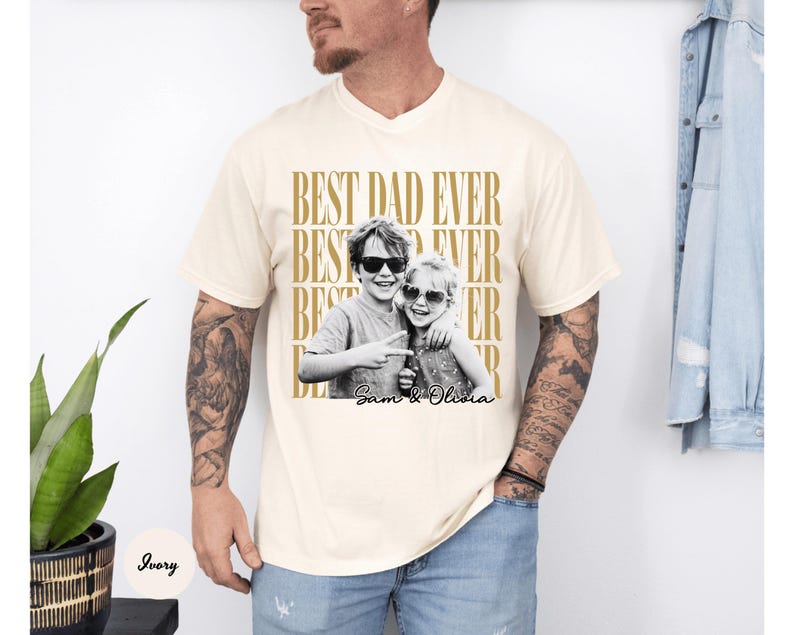 May include: Ivory-colored t-shirt with the text "BEST DAD EVER" in gold. A black and white photo of two children wearing sunglasses is centered on the shirt, with the names "Sam & Olivia" below.