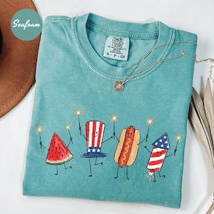 K&ouml;nnte beinhalten: T&uuml;rkisgr&uuml;nes Comfort Colors T-Shirt mit einer skurrilen Illustration einer Wassermelone, einem Uncle Sam Hut, einem Hotdog und einem Feuerwerk, die jeweils einen Wunderkerzenstab halten. Das Shirt hat einen runden Ausschnitt und kurze &Auml;rmel. Eine goldene Halskette liegt &uuml;ber dem Shirt.