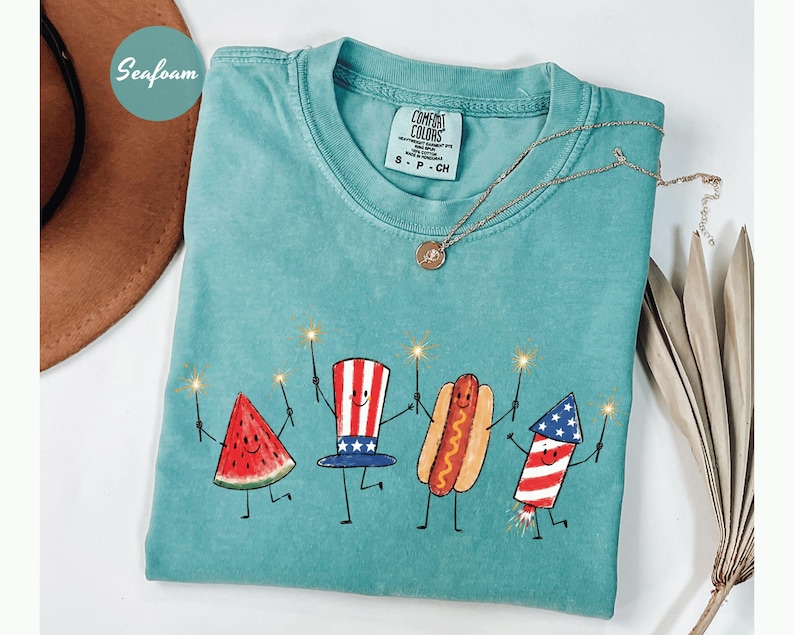 K&ouml;nnte beinhalten: T&uuml;rkisgr&uuml;nes Comfort Colors T-Shirt mit einer skurrilen Illustration einer Wassermelone, einem Uncle Sam Hut, einem Hotdog und einem Feuerwerk, die jeweils einen Wunderkerzenstab halten. Das Shirt hat einen runden Ausschnitt und kurze &Auml;rmel. Eine goldene Halskette liegt &uuml;ber dem Shirt.