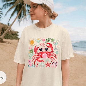 May include: Ivory-colored t-shirt featuring a whimsical beach-themed graphic. A red crab with heart-shaped eyes and a bubble gum bubble is surrounded by cocktails, seashells, and flip-flops. The shirt is a relaxed fit.