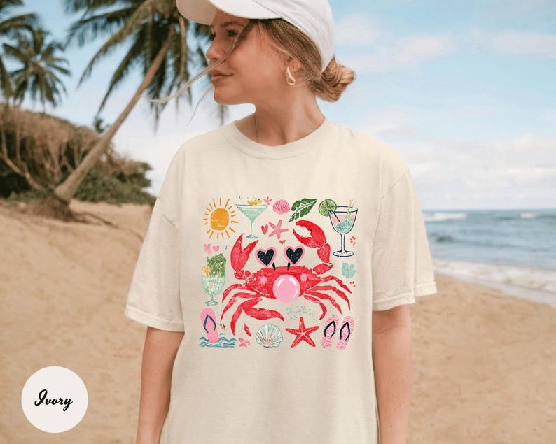 May include: Ivory-colored t-shirt featuring a whimsical beach-themed graphic. A red crab with heart-shaped eyes and a bubble gum bubble is surrounded by cocktails, seashells, and flip-flops. The shirt is a relaxed fit.