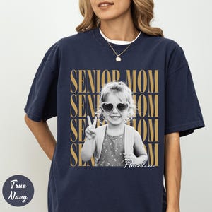 May include: A true navy short-sleeve t-shirt featuring a black and white photograph of a child wearing heart-shaped sunglasses and making a peace sign. The text SENIOR MOM is repeated in gold behind the photo, with the name Amelia in script.