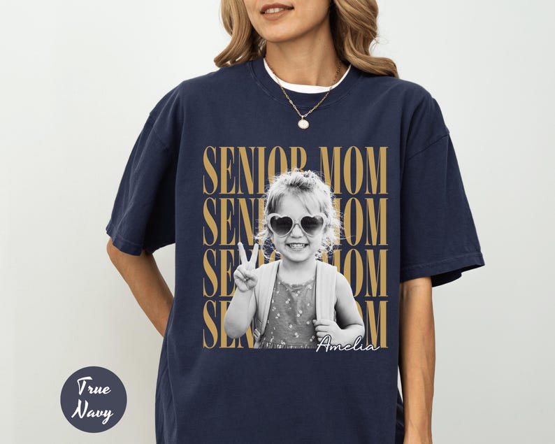 May include: A true navy short-sleeve t-shirt featuring a black and white photograph of a child wearing heart-shaped sunglasses and making a peace sign. The text SENIOR MOM is repeated in gold behind the photo, with the name Amelia in script.