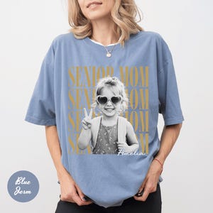 May include: A dusty blue short-sleeve t-shirt featuring a black and white photograph of a child wearing heart-shaped sunglasses and making a peace sign. The text SENIOR MOM is printed in large gold letters behind the photo, with the name Amelia below.