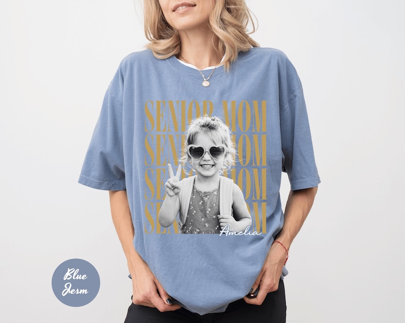 May include: A dusty blue short-sleeve t-shirt featuring a black and white photograph of a child wearing heart-shaped sunglasses and making a peace sign. The text SENIOR MOM is printed in large gold letters behind the photo, with the name Amelia below.
