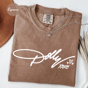 May include: An espresso-colored short-sleeve t-shirt featuring the white text Dolly xoxo with three small heart outlines. The shirt has a crew neckline and a Comfort Colors brand label on the inside collar. A silver chain necklace rests on top.