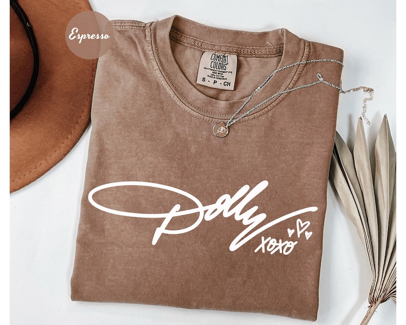 May include: An espresso-colored short-sleeve t-shirt featuring the white text Dolly xoxo with three small heart outlines. The shirt has a crew neckline and a Comfort Colors brand label on the inside collar. A silver chain necklace rests on top.