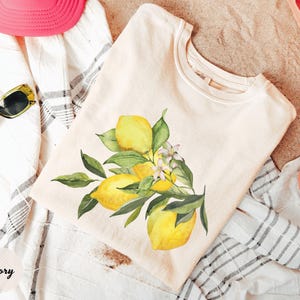 May include: Ivory-colored t-shirt featuring a watercolor illustration of lemons with green leaves and white flowers. The shirt is folded on a striped towel, with a pink hat and sunglasses visible. The word "Ivory" is printed on the bottom left.