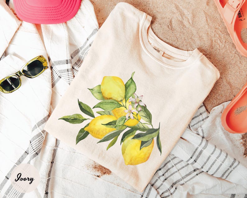 May include: Ivory-colored t-shirt featuring a watercolor illustration of lemons with green leaves and white flowers. The shirt is folded on a striped towel, with a pink hat and sunglasses visible. The word "Ivory" is printed on the bottom left.