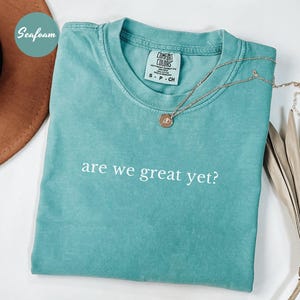 May include: Seafoam green Comfort Colors t-shirt with the phrase "are we great yet?" in white. A gold necklace with a pendant rests on the shirt. The shirt is folded neatly, and a brown hat is partially visible.