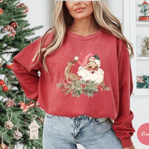May include: A red sweatshirt featuring a vintage-style Christmas design. The graphic includes Santa Claus, a reindeer, and festive greenery. The sweatshirt has long sleeves and a crew neck.