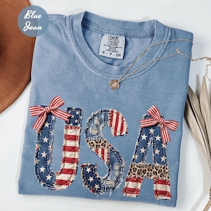 May include: Light blue t-shirt with the word "USA" appliqued in a patchwork design featuring stars, stripes, and leopard print. Red and white striped bows are attached to the letters. A gold necklace with a pendant is draped on the shirt.