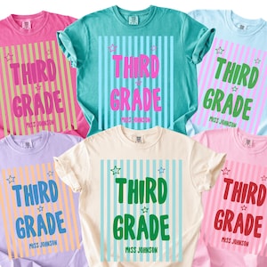 May include: A collection of six short-sleeved t-shirts in various pastel colours, each featuring a vertical striped background, small star icons, and the text "THIRD GRADE" followed by "MISS JOHNSON" printed in bold, colourful lettering.