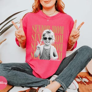 May include: A bright pink short-sleeve t-shirt featuring a black and white photograph of a young child wearing heart-shaped sunglasses and making a peace sign. The text SENIOR MOM is repeated in gold behind the photo, with the name Amelia below.