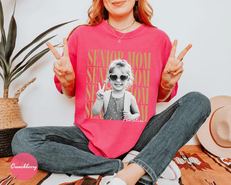 May include: A bright pink short-sleeve t-shirt featuring a black and white photograph of a young child wearing heart-shaped sunglasses and making a peace sign. The text SENIOR MOM is repeated in gold behind the photo, with the name Amelia below.