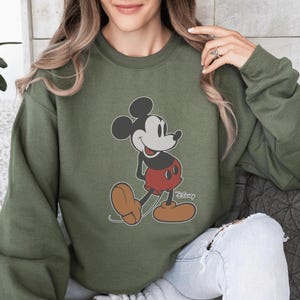 May include: An olive green sweatshirt with a Mickey Mouse graphic. The cartoon character is shown in his classic pose, wearing red shorts, brown shoes, and white gloves. The Disney logo is printed on the bottom right of the sweatshirt.
