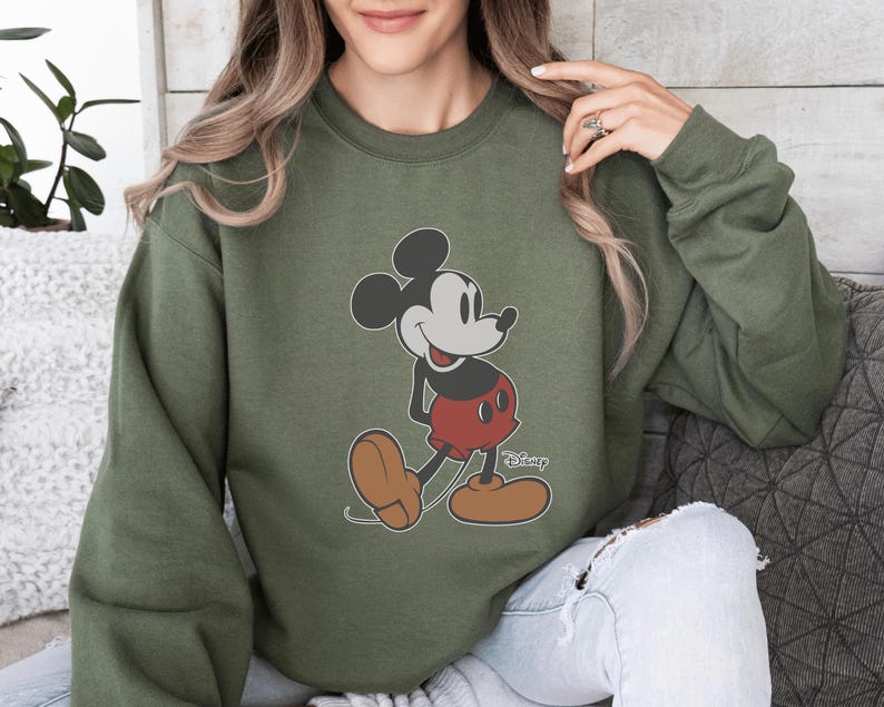 May include: An olive green sweatshirt with a Mickey Mouse graphic. The cartoon character is shown in his classic pose, wearing red shorts, brown shoes, and white gloves. The Disney logo is printed on the bottom right of the sweatshirt.