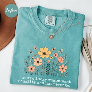 Peut inclure: T-shirt vert d'eau avec un motif floral composé de fleurs jaunes et pêche, de papillons et le texte "You're lucky women want equality and not revenge." Un collier en or repose sur le t-shirt.