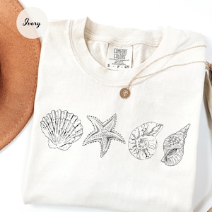 May include: Ivory-colored t-shirt featuring a black and white line drawing of a seashell, starfish, and two other shells. The shirt has a crew neck and short sleeves. A gold necklace with a pendant is draped across the shirt.