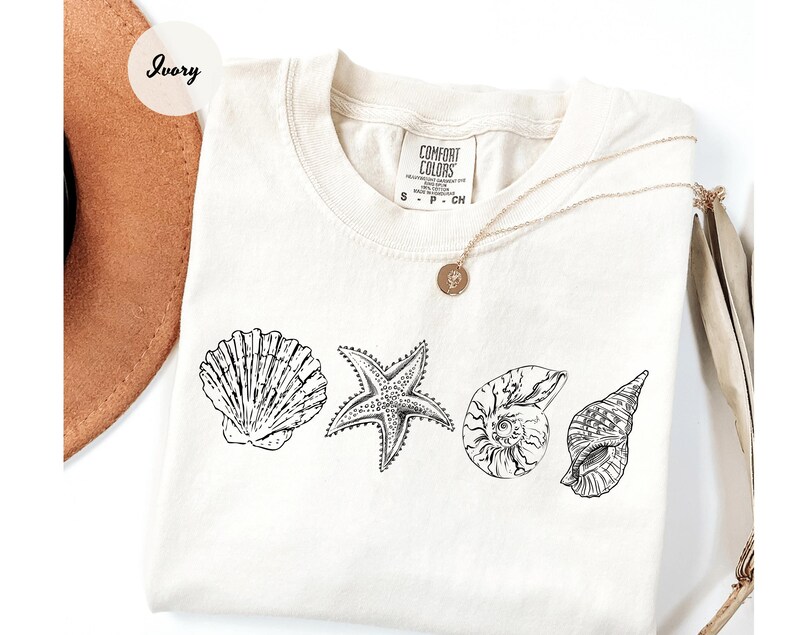 May include: Ivory-colored t-shirt featuring a black and white line drawing of a seashell, starfish, and two other shells. The shirt has a crew neck and short sleeves. A gold necklace with a pendant is draped across the shirt.
