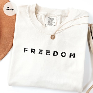 May include: Ivory-colored t-shirt with the word "FREEDOM" printed in black block letters. A gold necklace with a pendant rests on the shirt. A brown hat is partially visible in the upper left corner.