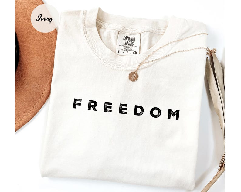 May include: Ivory-colored t-shirt with the word "FREEDOM" printed in black block letters. A gold necklace with a pendant rests on the shirt. A brown hat is partially visible in the upper left corner.