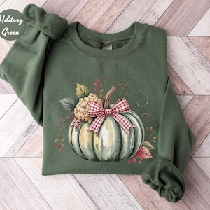 May include: Military green sweatshirt featuring a watercolor design of a pumpkin with a red and white checkered bow, surrounded by fall foliage. The pumpkin is light green with white highlights. The sweatshirt has a crew neck and long sleeves.