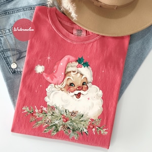 May include: A red t-shirt featuring a vintage Santa Claus graphic with a pink hat and white beard. The Santa Claus graphic is surrounded by greenery and berries. The shirt is folded on top of blue jeans.