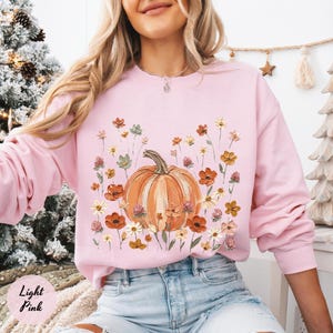 May include: A light pink long-sleeve sweatshirt featuring a central graphic of an orange pumpkin surrounded by various colorful wildflowers in shades of red, yellow, and green. The crewneck garment has a ribbed collar, cuffs, and hem.