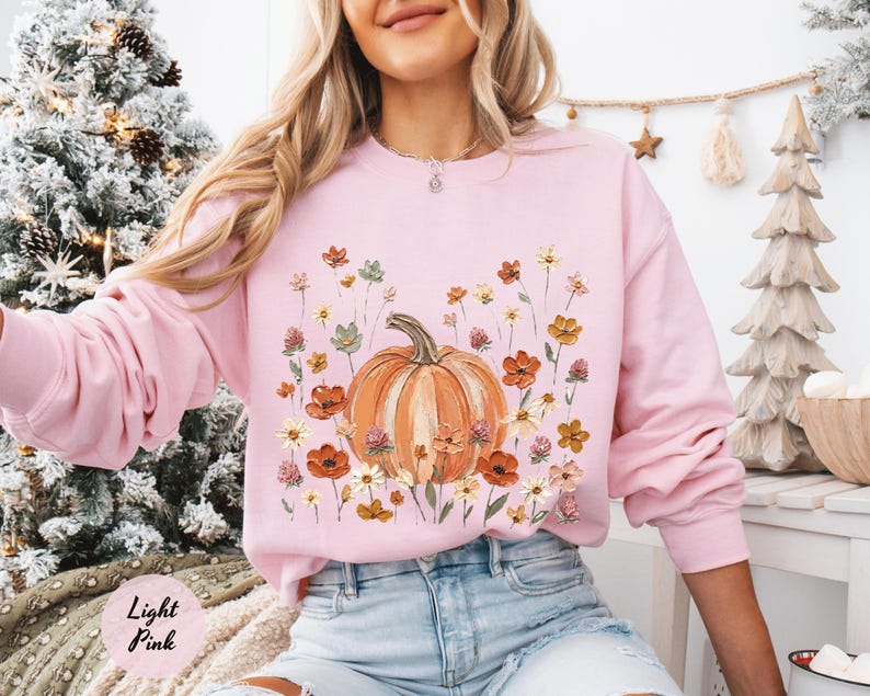 May include: A light pink long-sleeve sweatshirt featuring a central graphic of an orange pumpkin surrounded by various colorful wildflowers in shades of red, yellow, and green. The crewneck garment has a ribbed collar, cuffs, and hem.